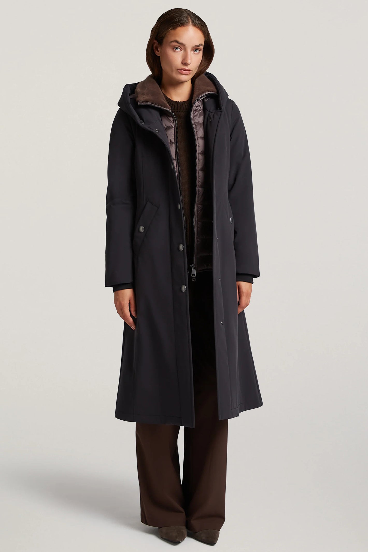 Woman wearing a long black coat with a hood on a light gray background