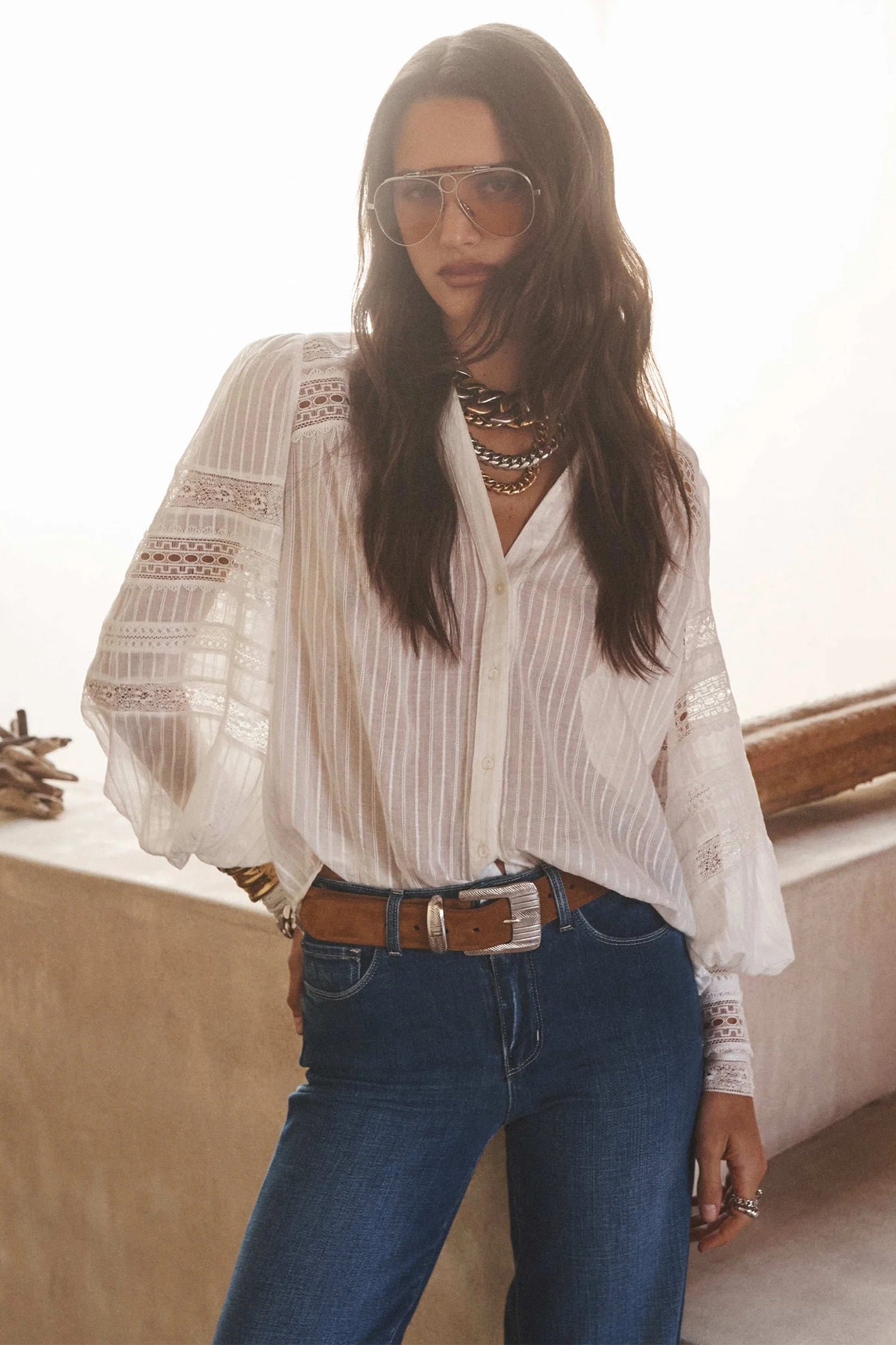 Woman wearing a white blouse with lace details and blue jeans, standing indoors.