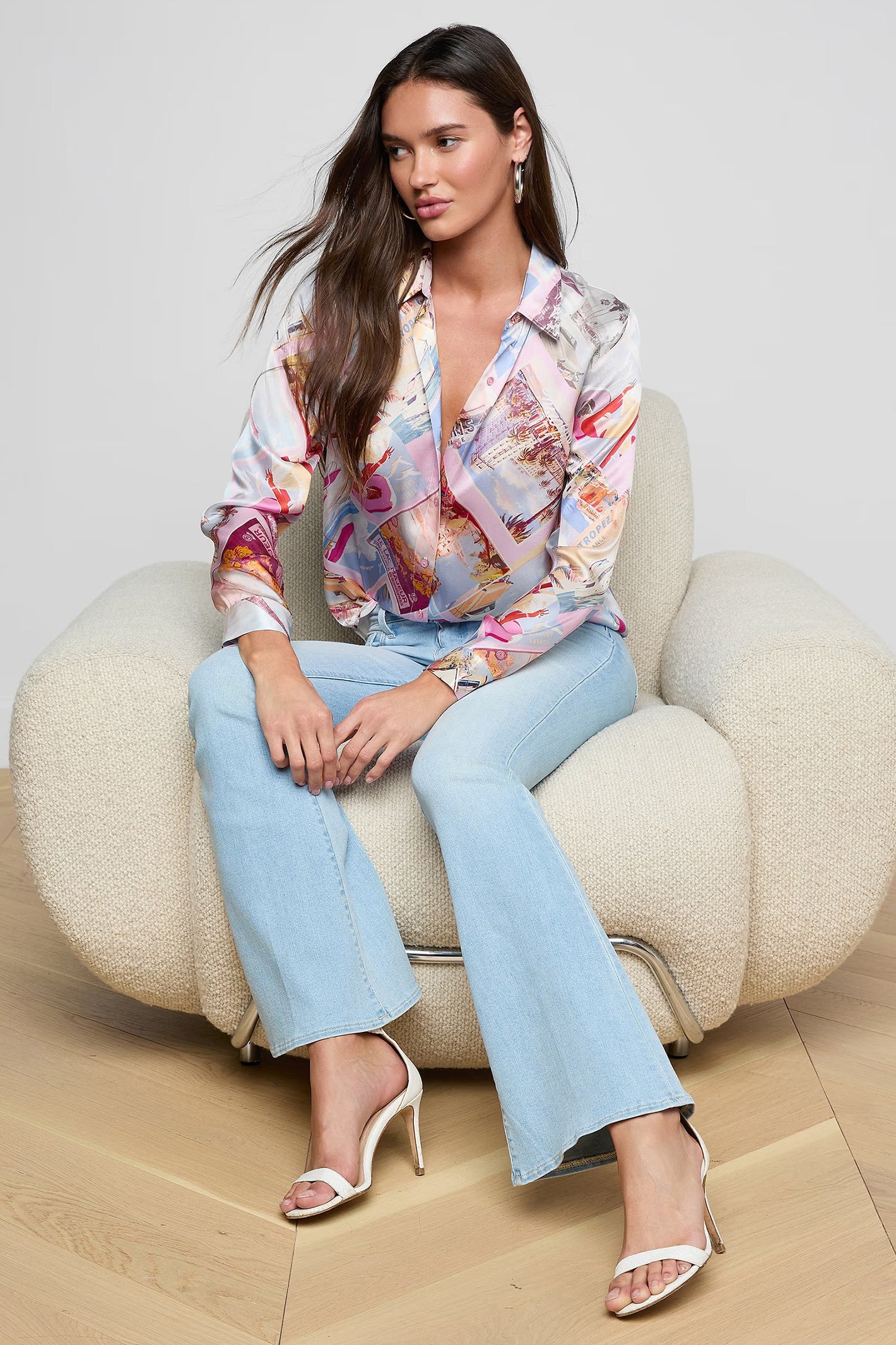 Woman sitting on a beige chair wearing a colorful blouse and light blue jeans.