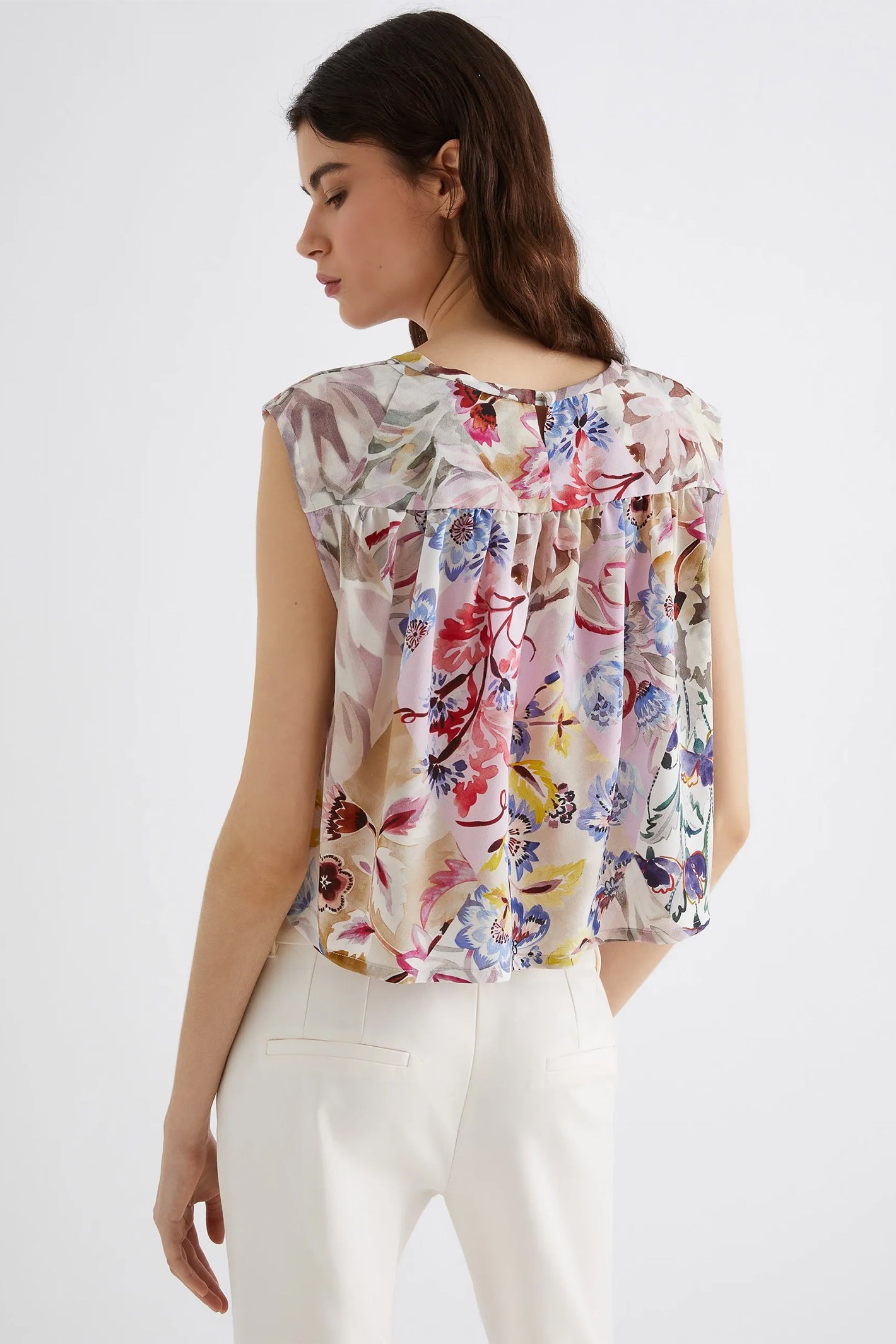 Woman wearing a sleeveless floral top and white pants on a plain background