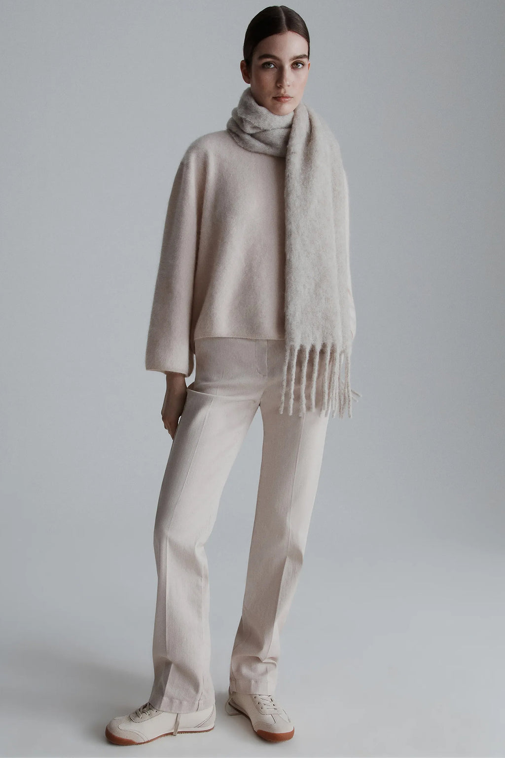 Person wearing a beige sweater, scarf, and pants against a plain background