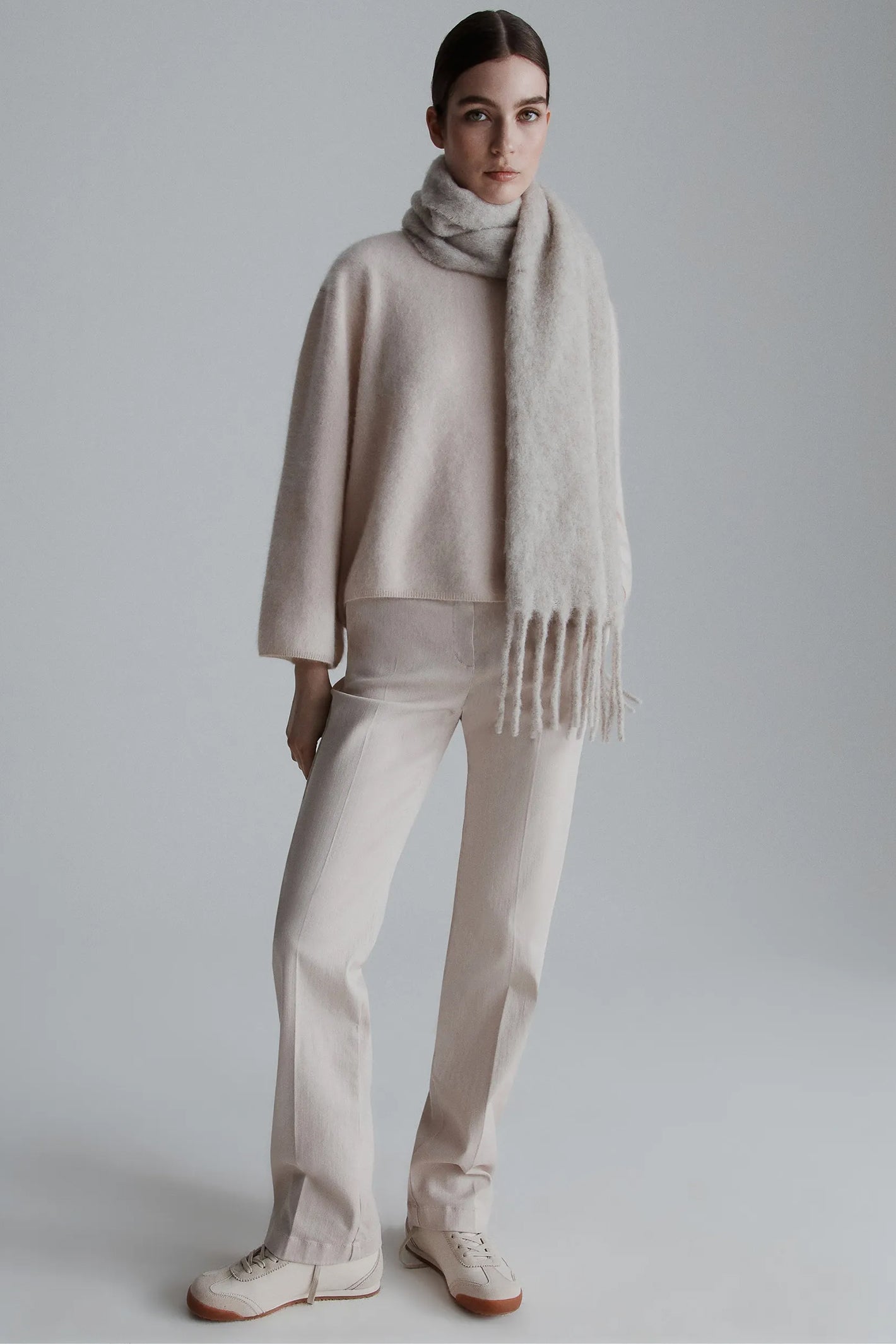 Person wearing a beige sweater, scarf, and pants against a plain background