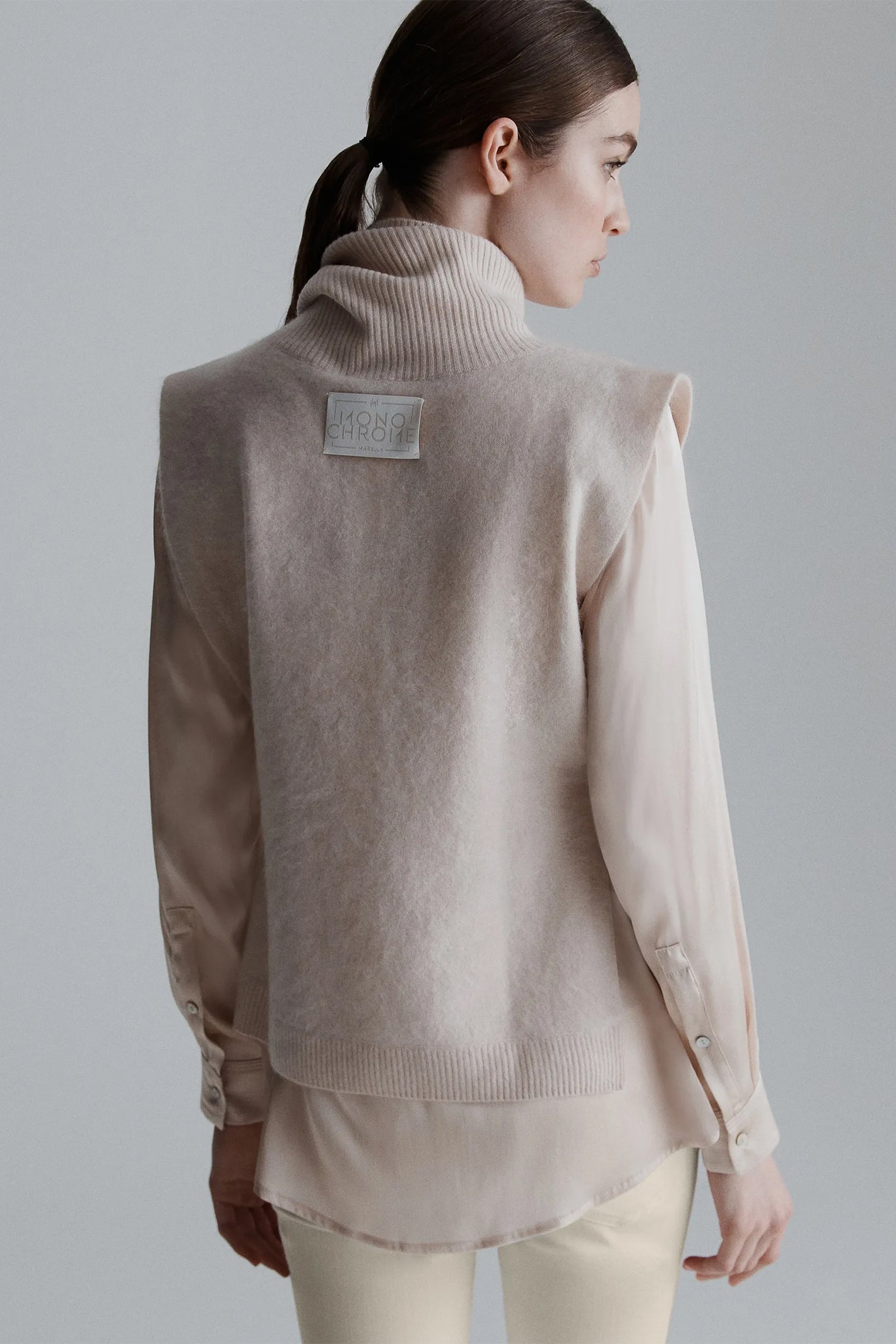 Woman wearing a beige sweater with a visible brand label on a plain background