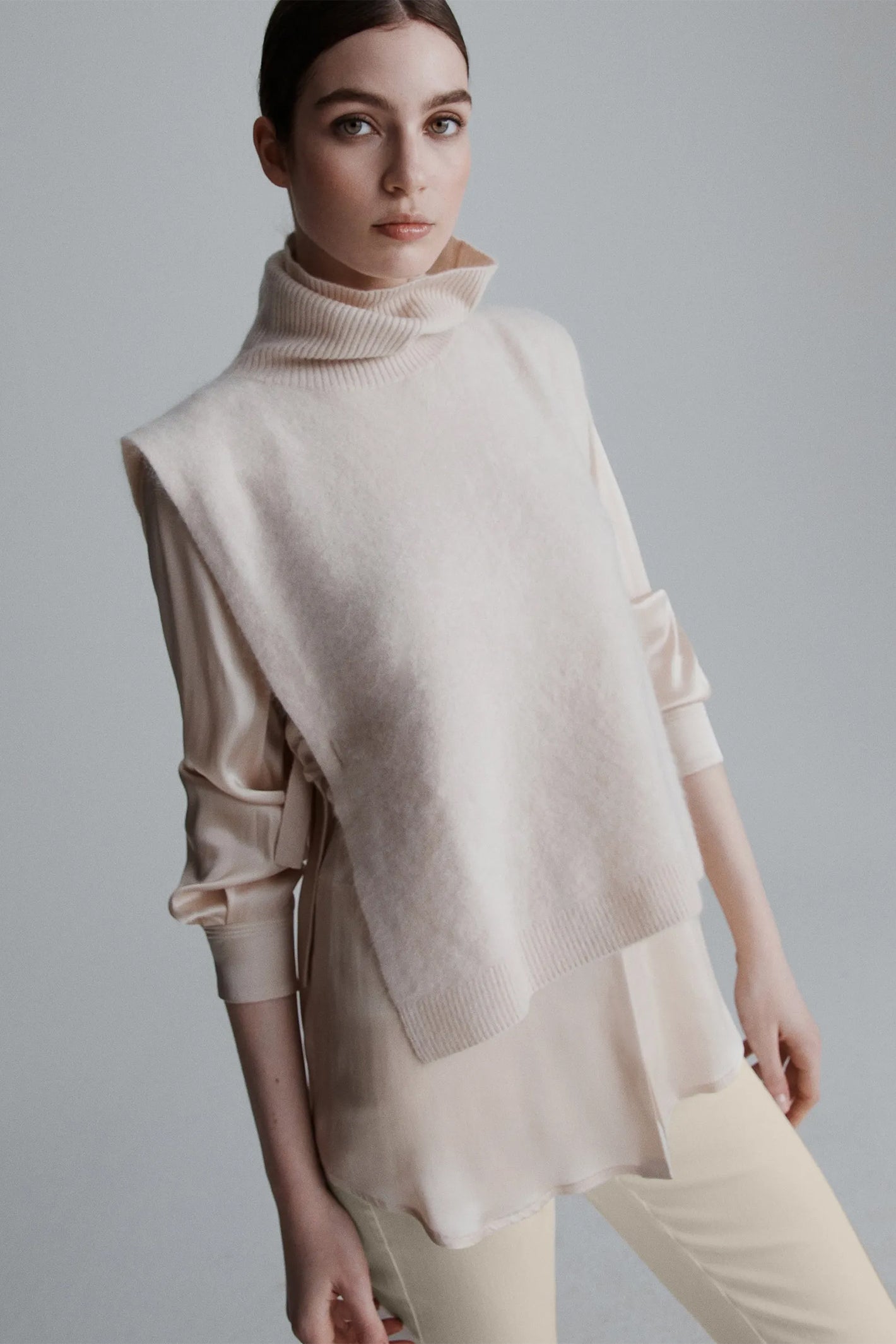 Woman wearing a beige turtleneck sweater with a cape over a plain background