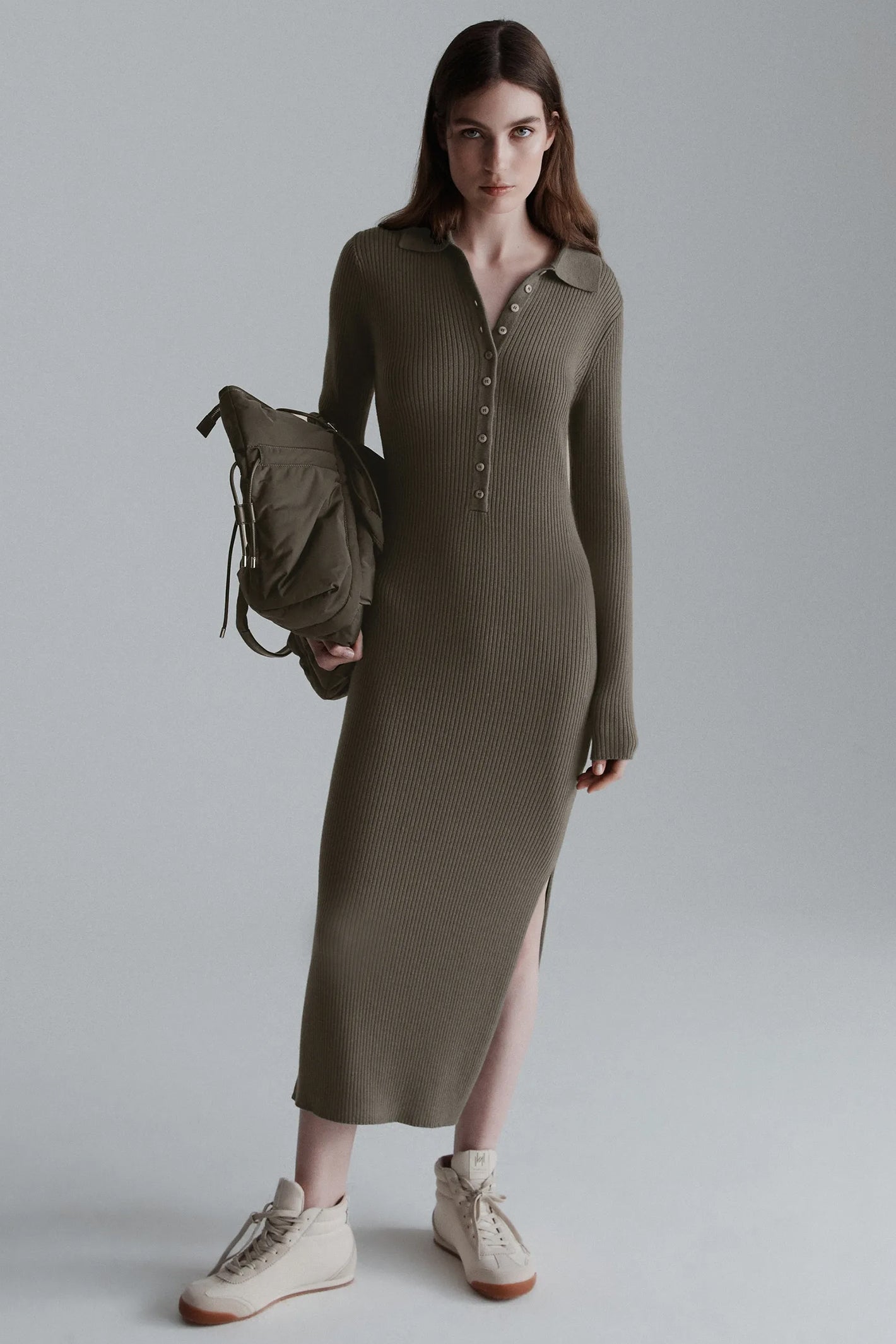 Woman wearing a long-sleeve olive green dress holding a backpack against a plain background
