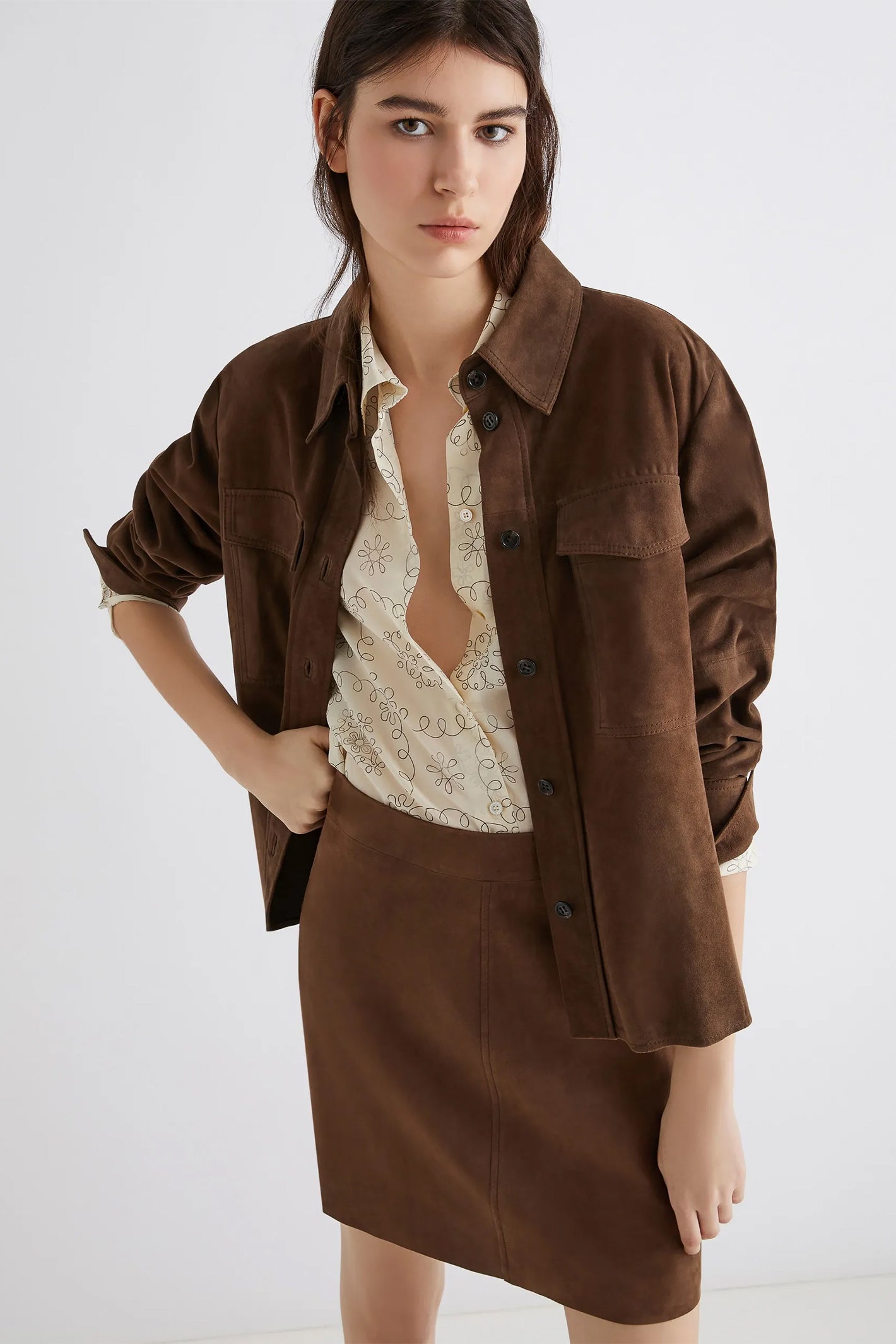 Woman wearing a brown jacket over a lace top and skirt on a white background