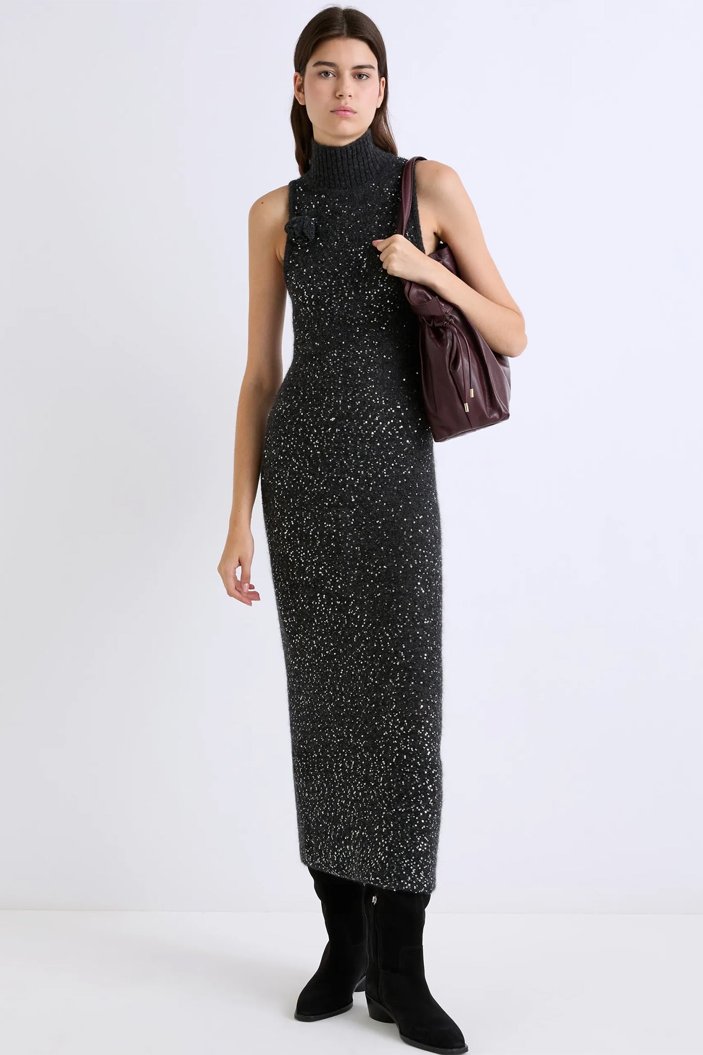 Woman wearing a grey sleeveless dress with a high neckline, holding a brown handbag on a white background.