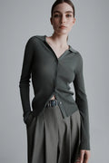 Woman wearing a green ribbed cardigan over a black shirt with a belt, standing against a gray background