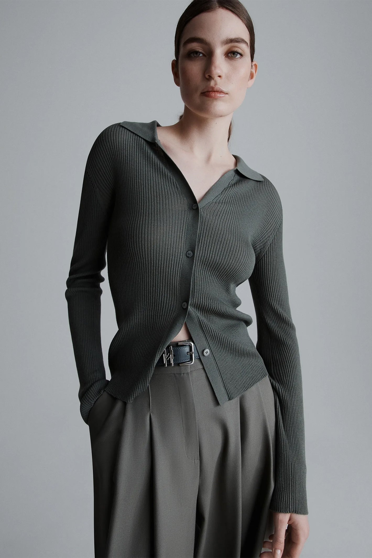 Woman wearing a green ribbed cardigan over a black shirt with a belt, standing against a gray background