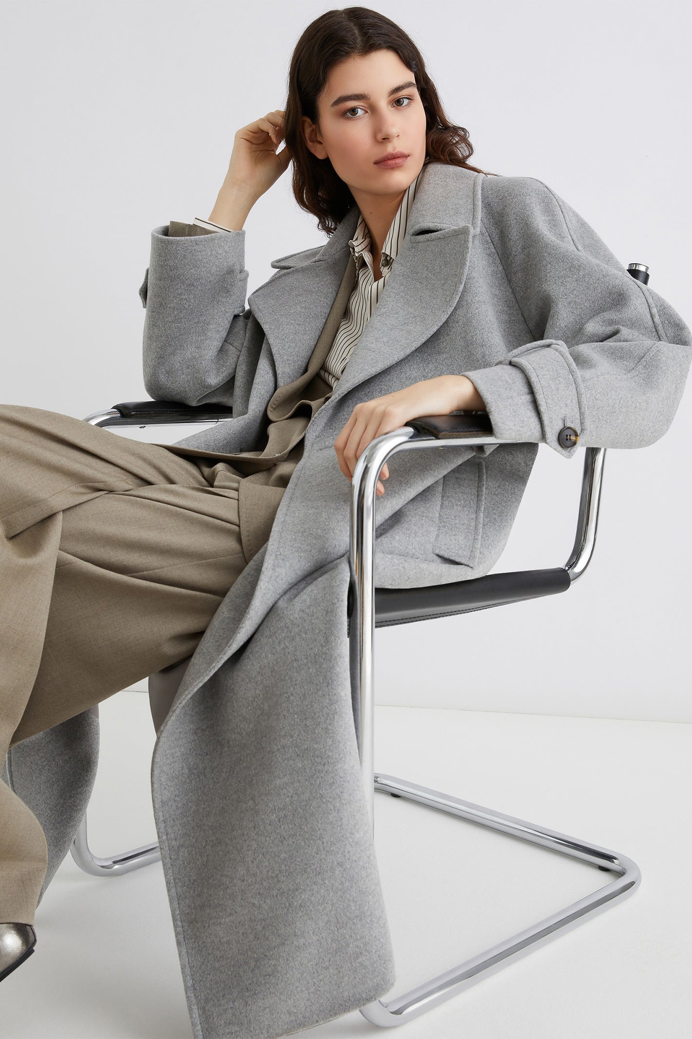 Woman wearing a long gray coat sitting on a chair against a white background