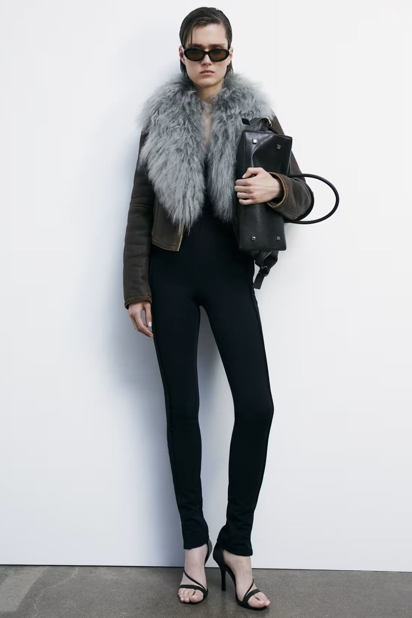 Person wearing a dark jacket with fur collar, black leggings, and sunglasses against a white background