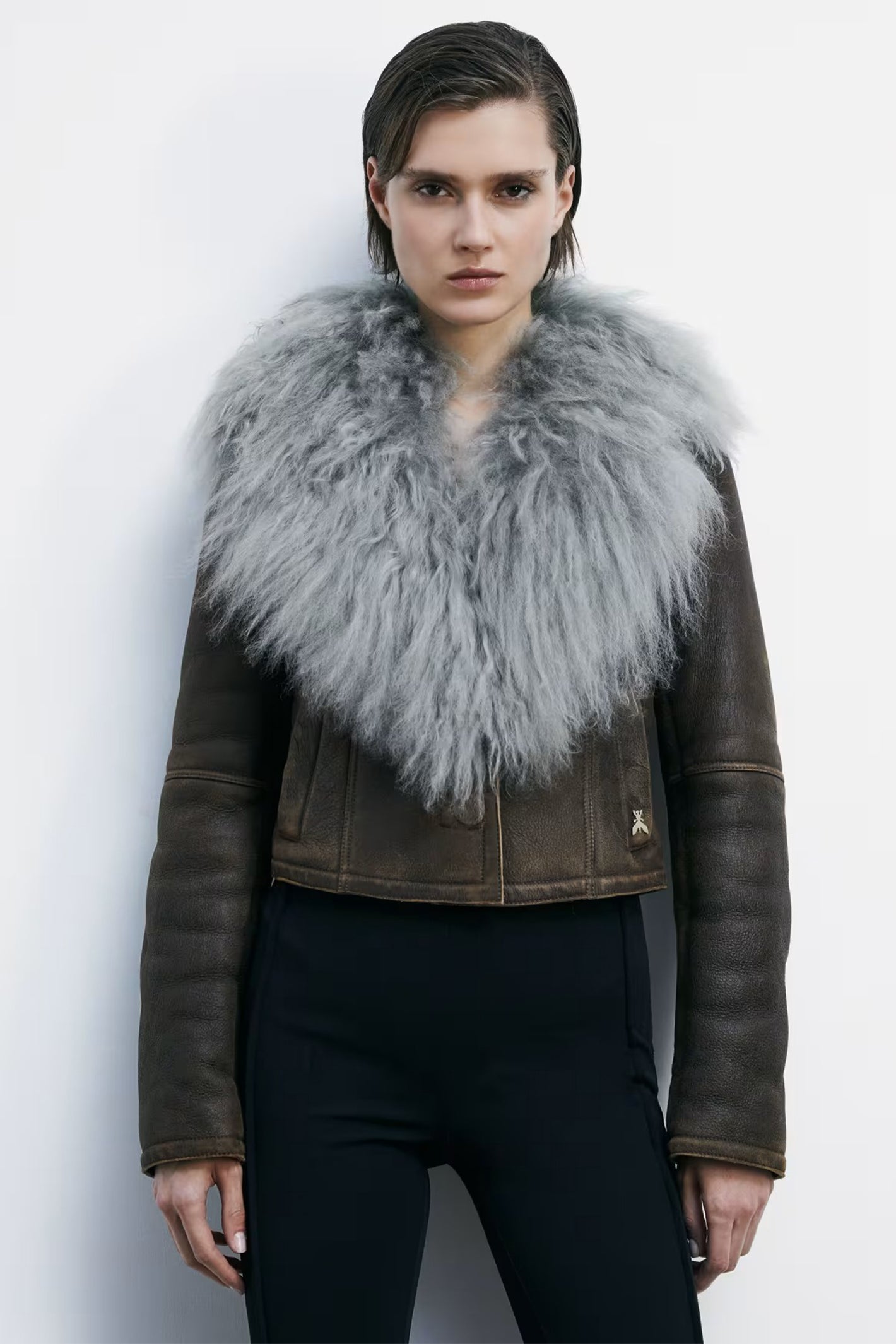Person wearing a brown leather jacket with a large gray fur collar against a white background