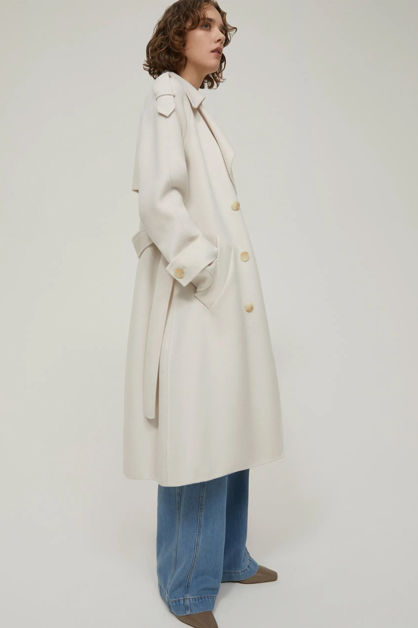 Person wearing a long beige coat and blue jeans on a white background