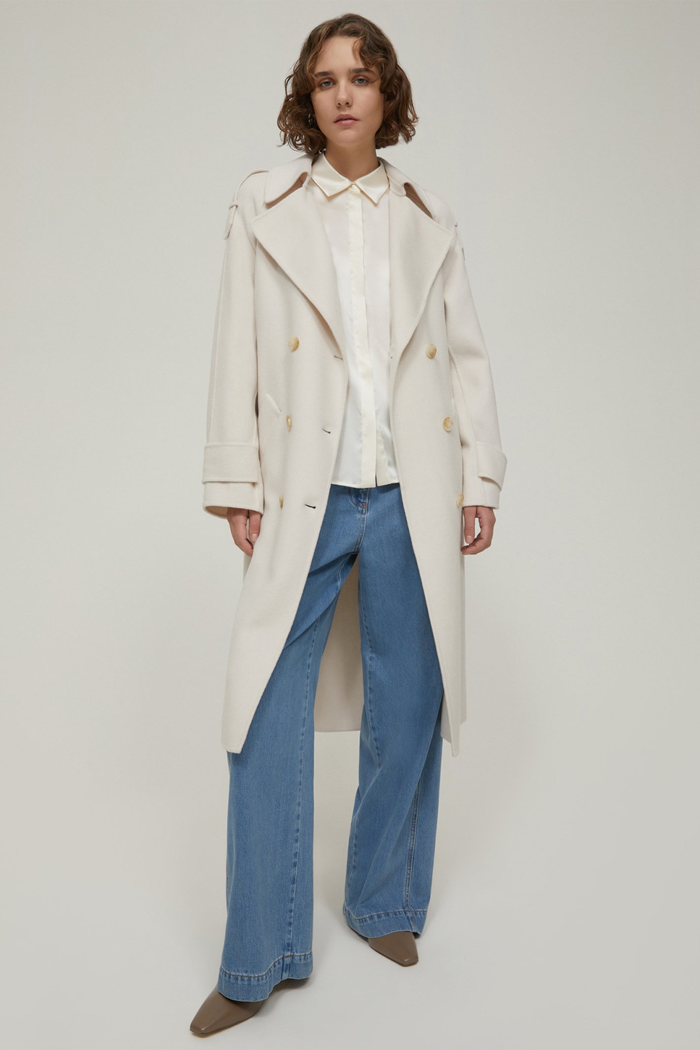 Person wearing a long cream coat and blue jeans on a white background